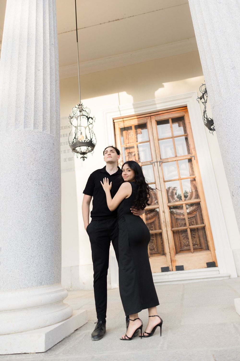 A couple in matching black outfits pose elegantly against white columns and ornate architecture.
