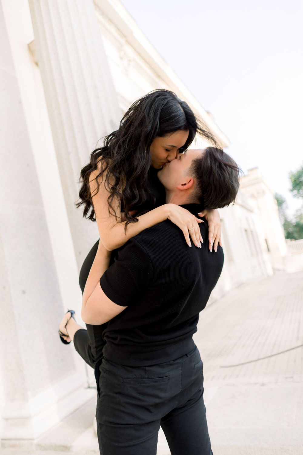 A romantic couple embraces against white columns, with the woman wearing black being lifted by her partner.