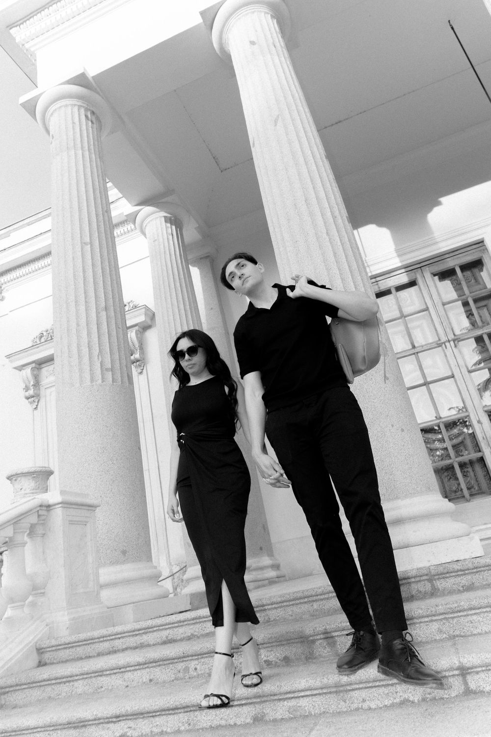 Two people in black outfits pose on steps with classical white columns in a dramatic black and white composition.