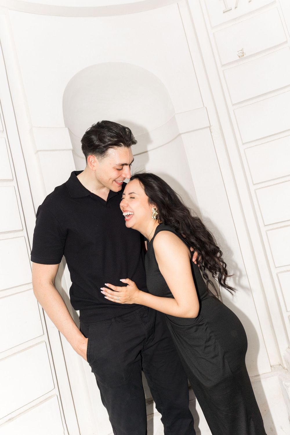 A couple dressed in matching black outfits share a candid laughing moment against a white wall with archway.