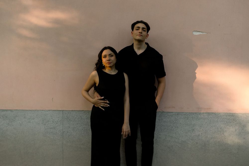 Two people dressed in black stand against a wall beneath a cloudy sky at dusk.
