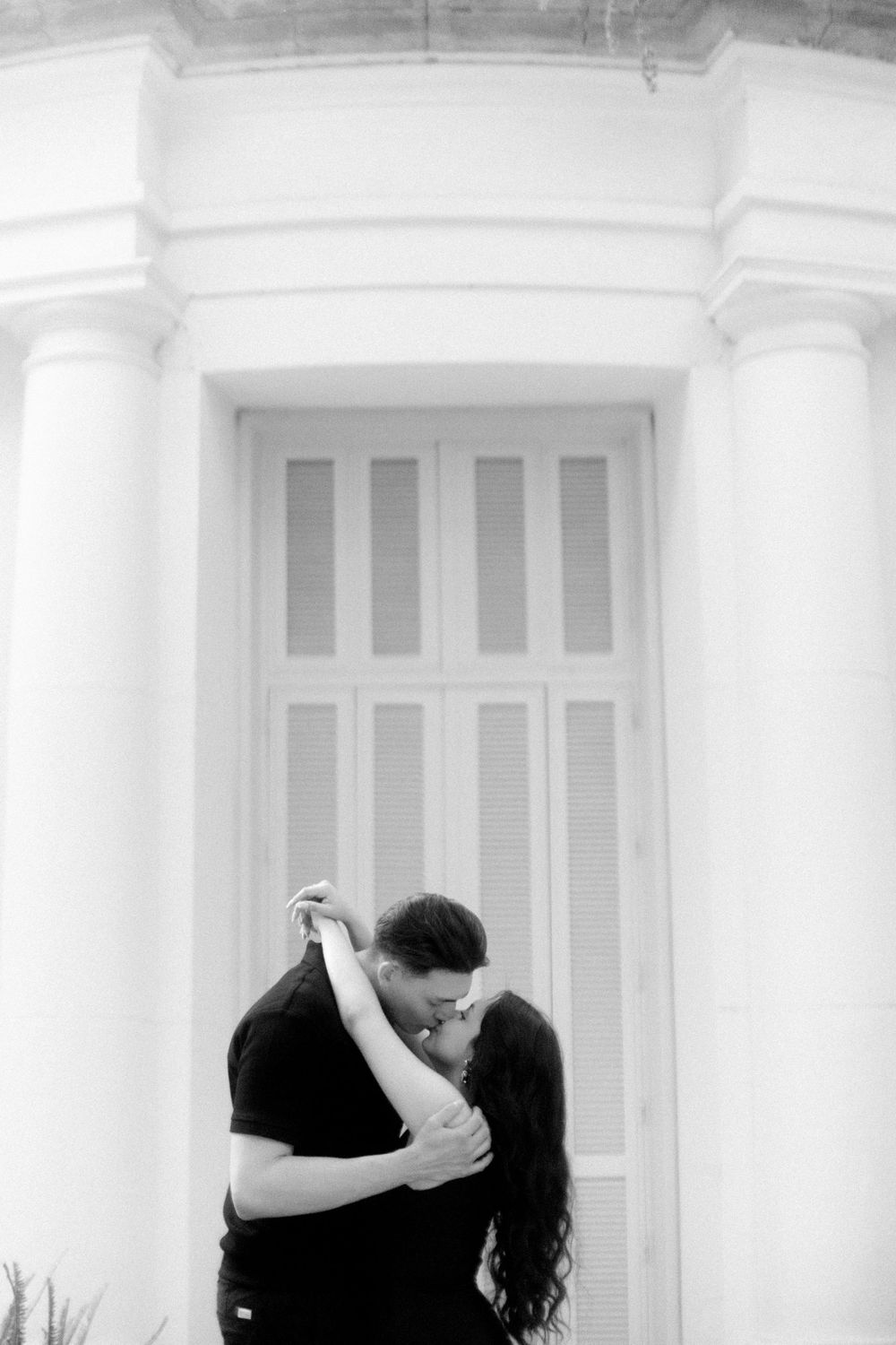 A romantic black and white photo of a couple sharing an intimate embrace in front of a white columned doorway.