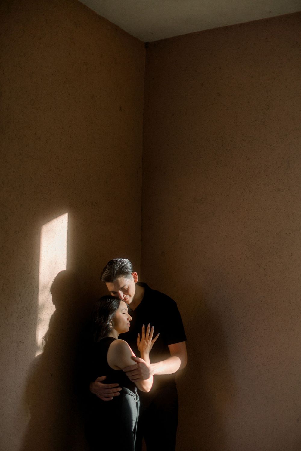 A couple embraces in a minimalist room with dramatic window light casting shadows on brown walls.
