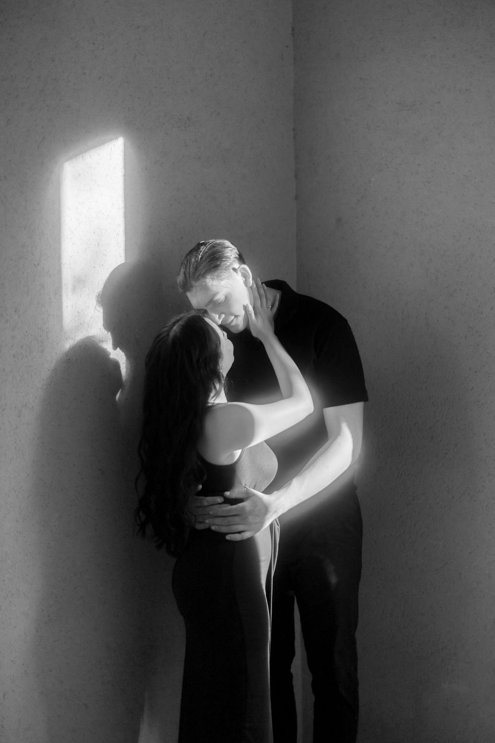 Intimate black and white photograph of a couple embracing in shadows with dramatic window lighting.