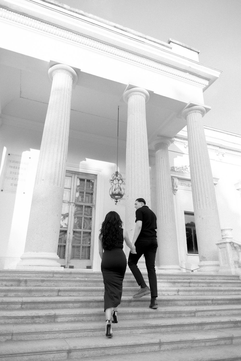 A couple ascends white marble steps leading to a grand columned entrance of a classical building in black and white.