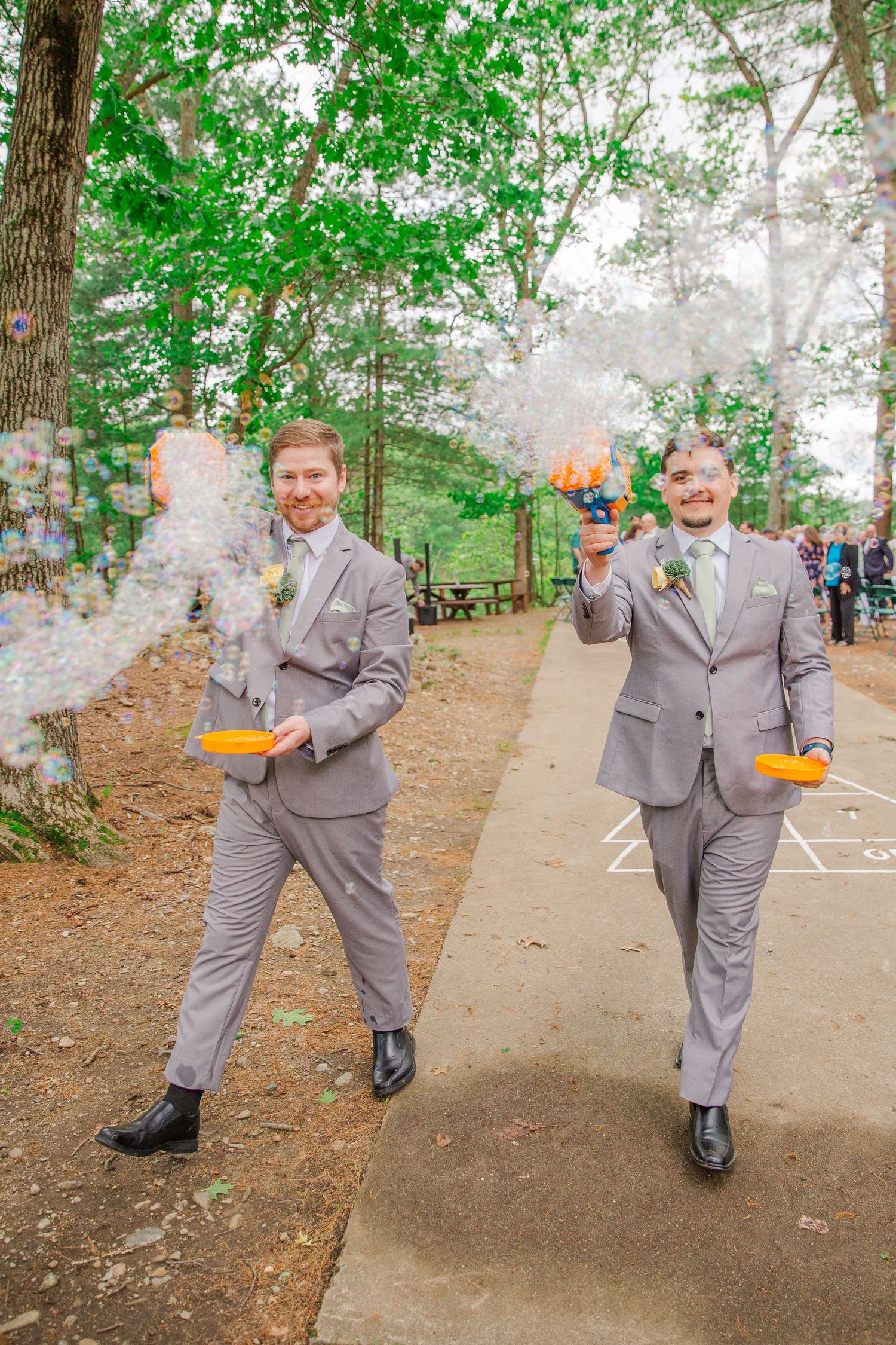 Two people in matching gray suits walk on a path in a wooded area while holding bubbling beverage glasses.