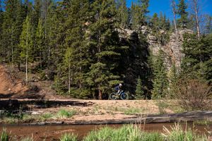 Cyclist jumps over stream near rocky cliff face surrounded by evergreen forest.