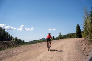 Cyclist rides along remote dirt road under bright blue sky in mountain landscape.