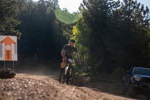 Mountain biker rides on dusty dirt trail through forested terrain on a sunny day.