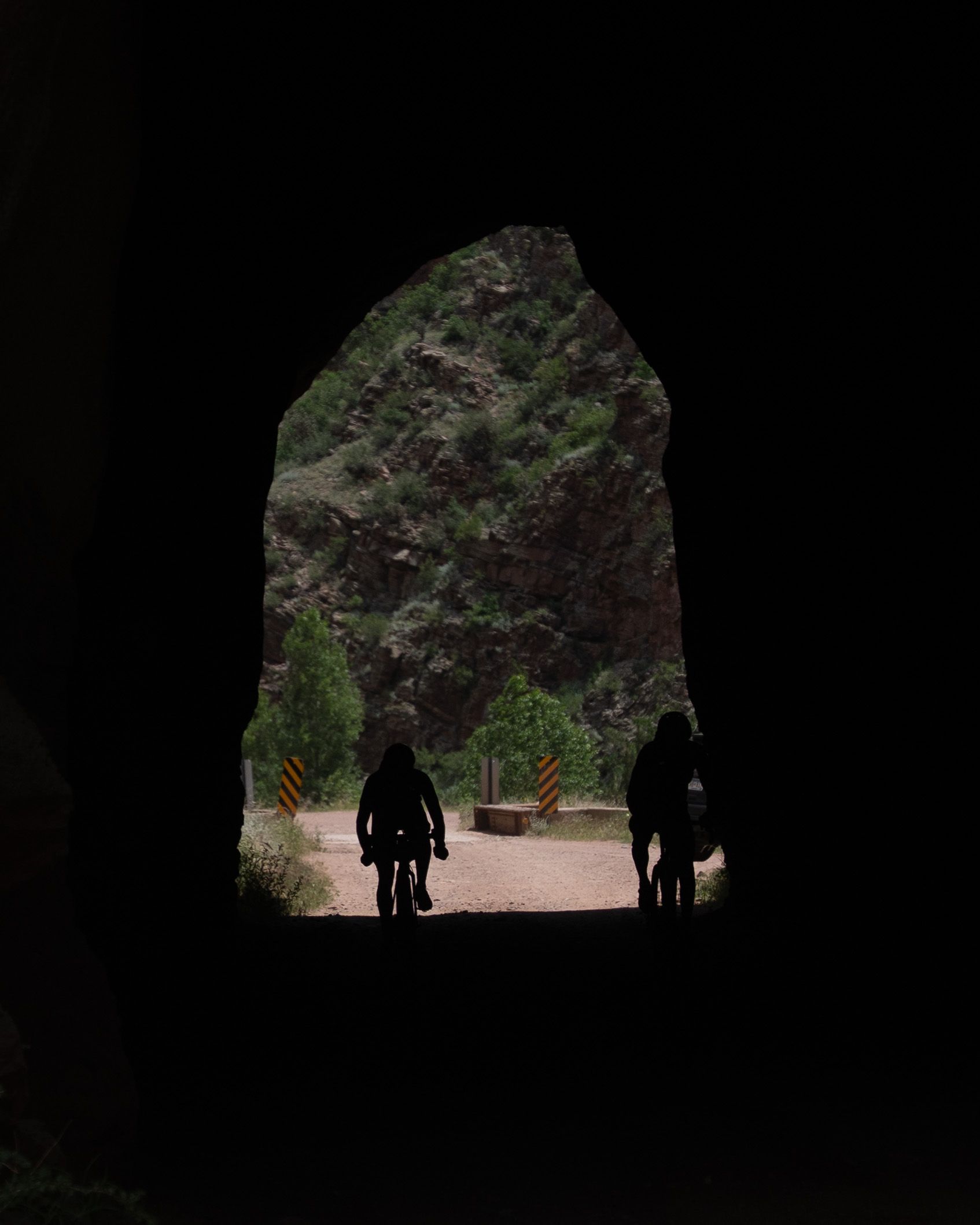 Dark tunnel opening with backlit silhouettes of hikers entering through a natural stone archway.