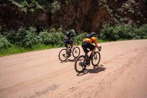 A group of cyclists navigate a rugged dirt trail through a mountainous forest landscape.