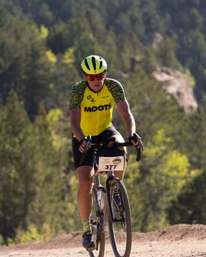 Cyclist in yellow jersey rides mountain bike on dirt trail surrounded by mountain terrain and forest.