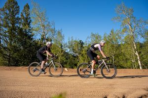 Two cyclists ride mountain bikes on a dirt trail through a scenic wooded area on a sunny day.