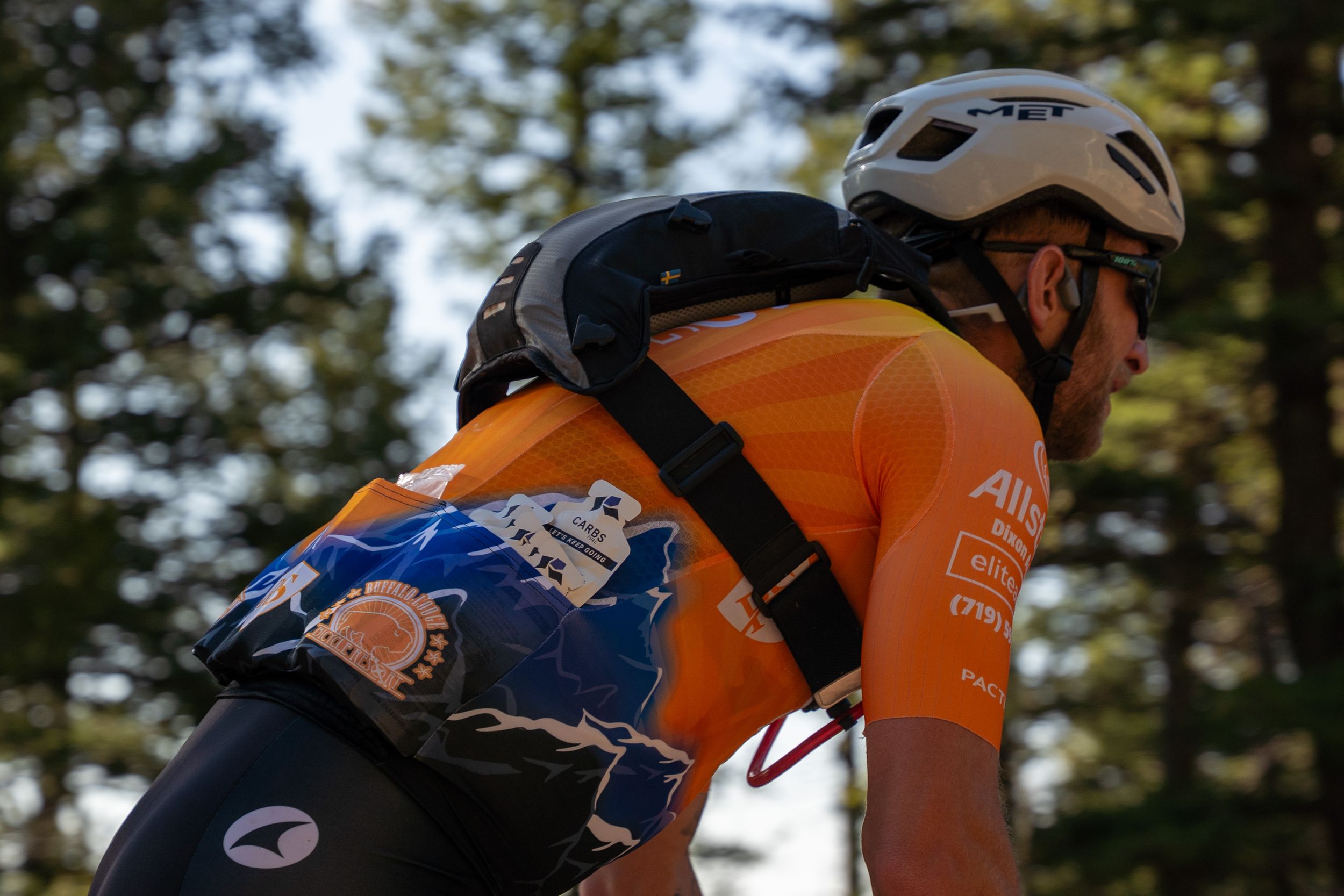 A cyclist in an orange jersey and white helmet rides through a wooded area on a sunny day.