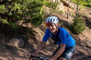 Mountain biker wearing blue jersey and helmet rides on dirt trail through wooded terrain.