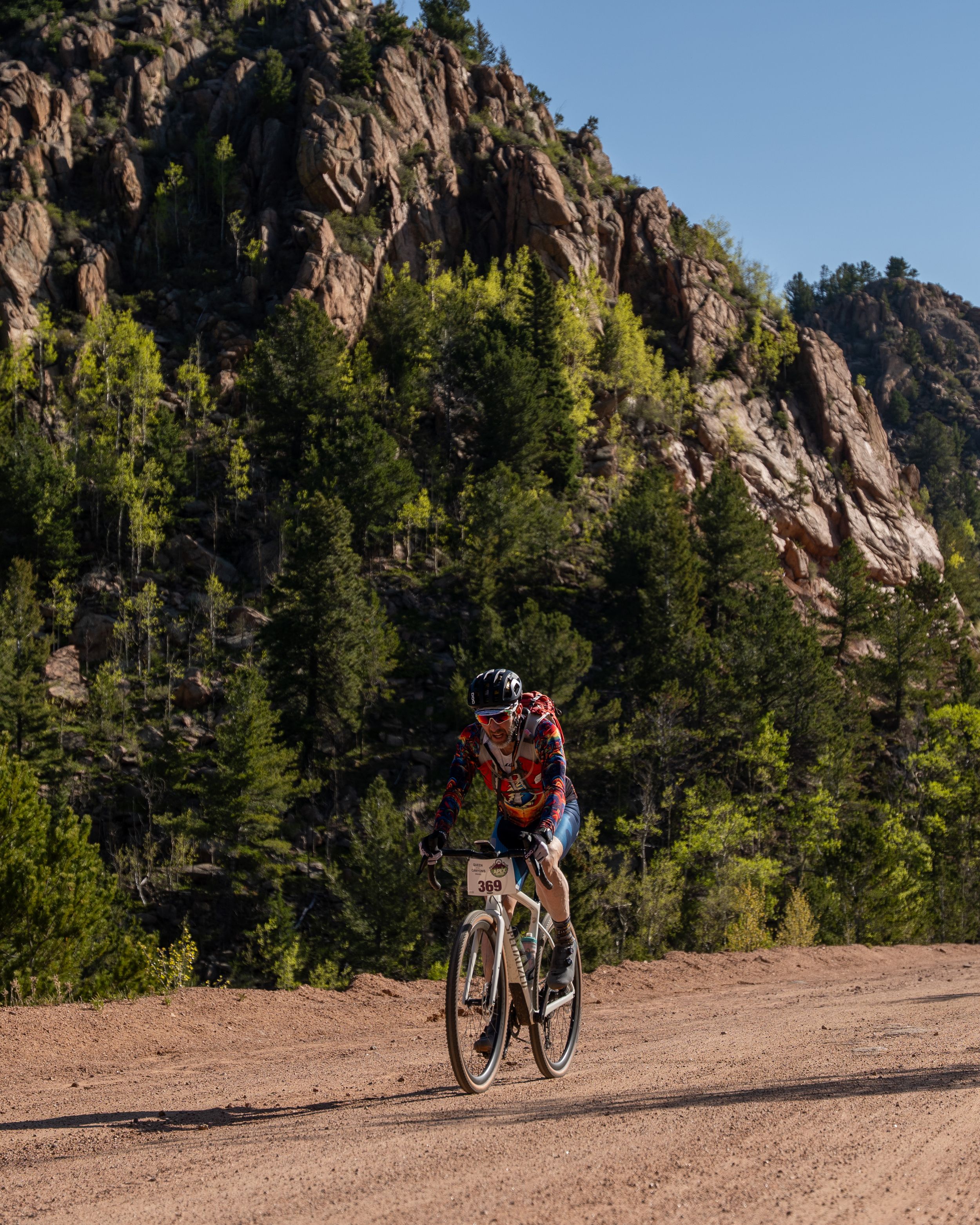 Mountain biker navigates a scenic trail with rocky cliffs and evergreen trees in the background.
