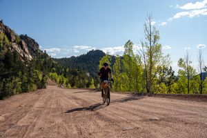 A cyclist rides on a dirt road through mountainous terrain with green trees and blue skies in the background.