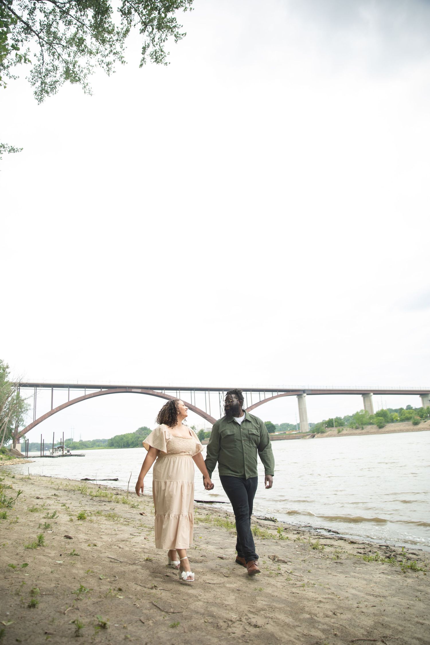 Couple walks hand in hand along riverbank with scenic arched bridge in background on cloudy day.