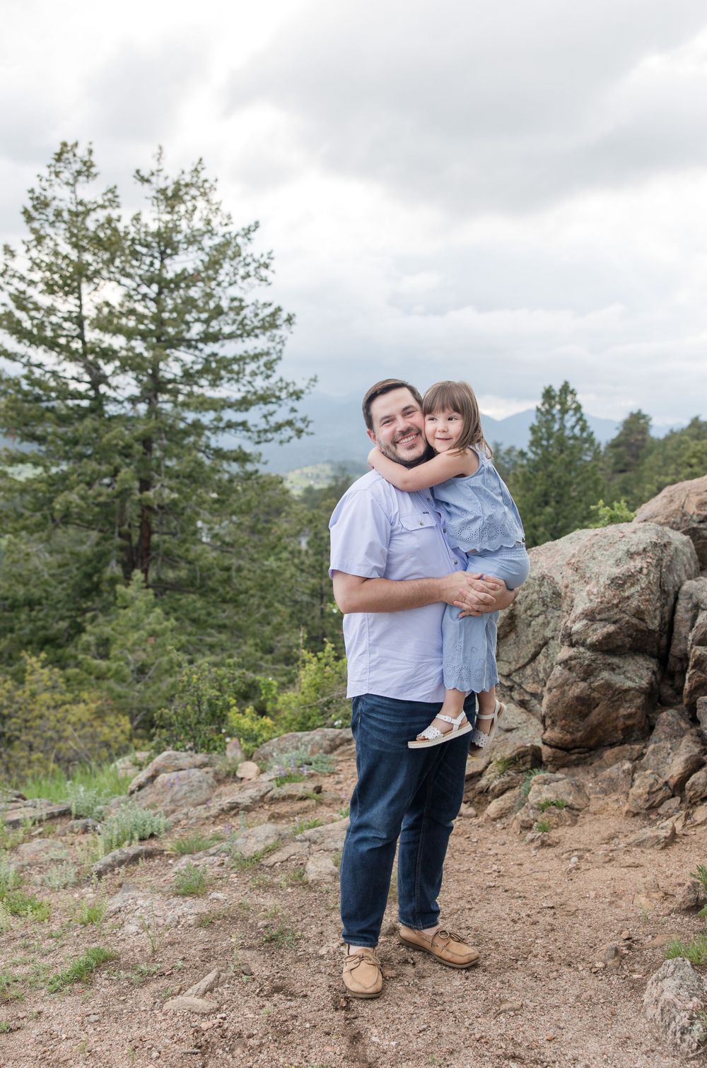 A parent and child share a tender moment while standing on a rocky overlook surrounded by pine trees.