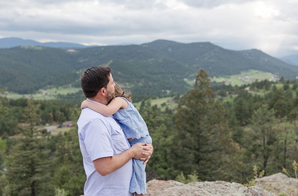 A scenic mountain view serves as backdrop for an embrace on a rocky viewpoint among evergreen trees.