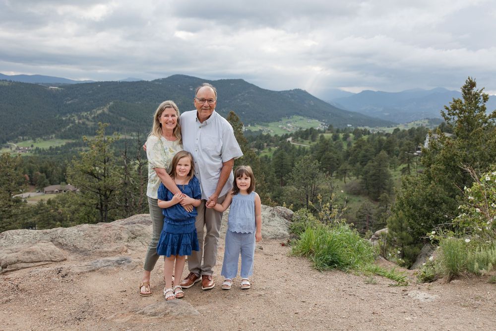 A group poses together on a rocky mountain overlook with evergreen trees and mountains in the background.