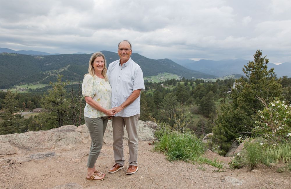 A couple poses together on a mountain trail overlook with sweeping mountain vistas behind them.