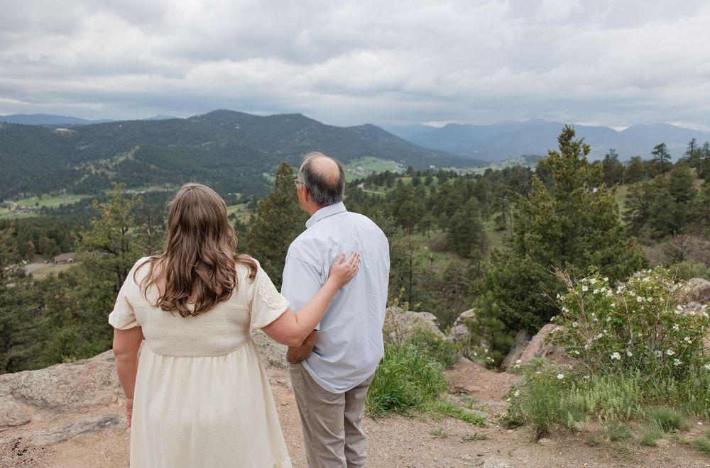 Two people standing on a mountain overlook admiring the sweeping vista of forested peaks under cloudy skies.