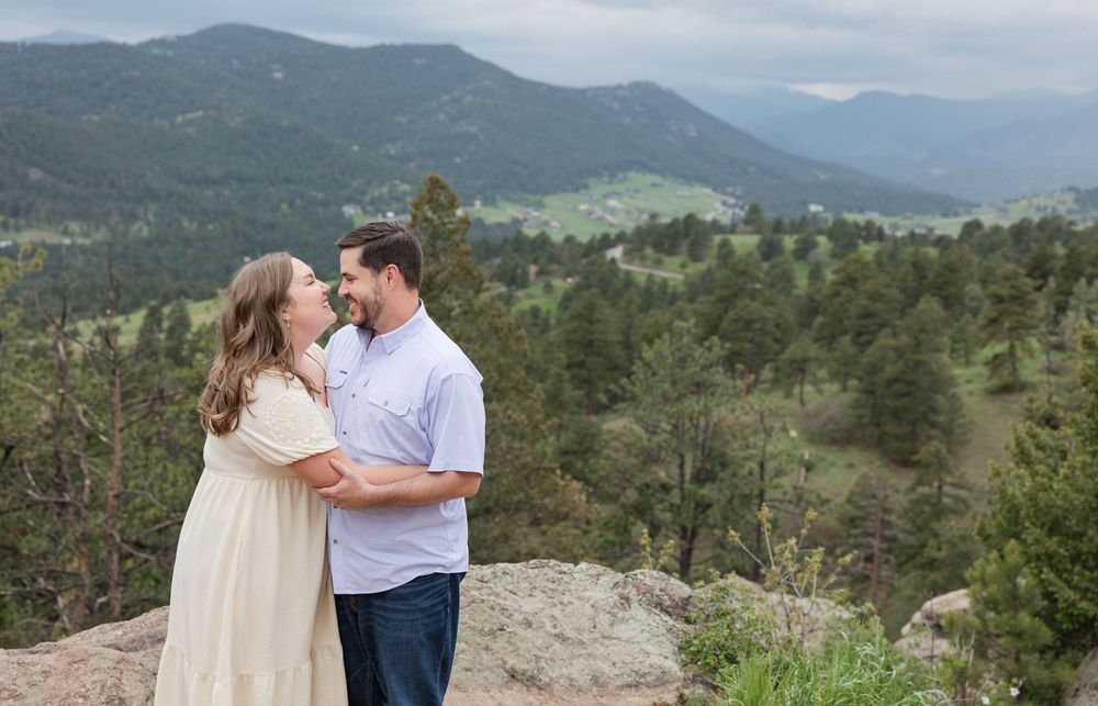 A romantic couple embracing on a scenic mountain overlook with evergreen forest and mountains in the distance.