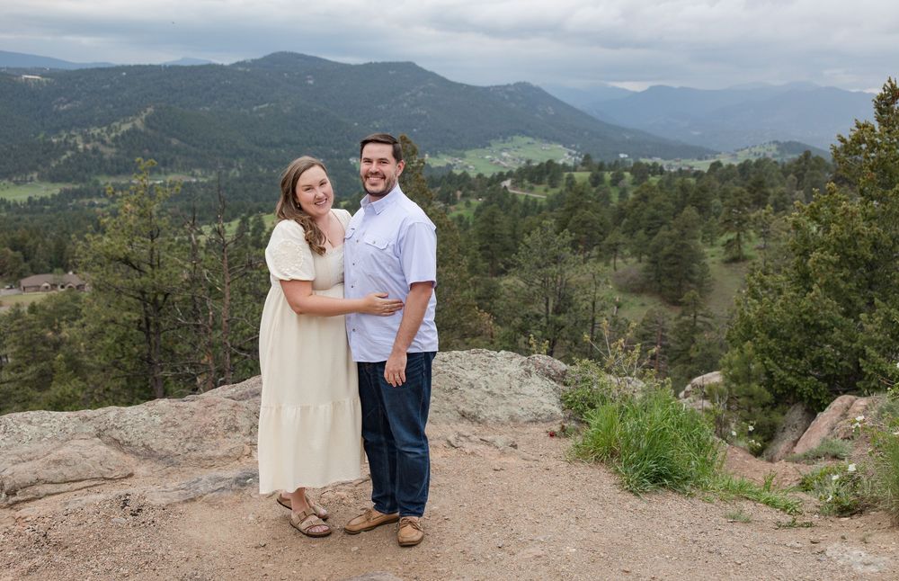 A couple poses together on a scenic mountain trail with rolling hills and forests in the distance.