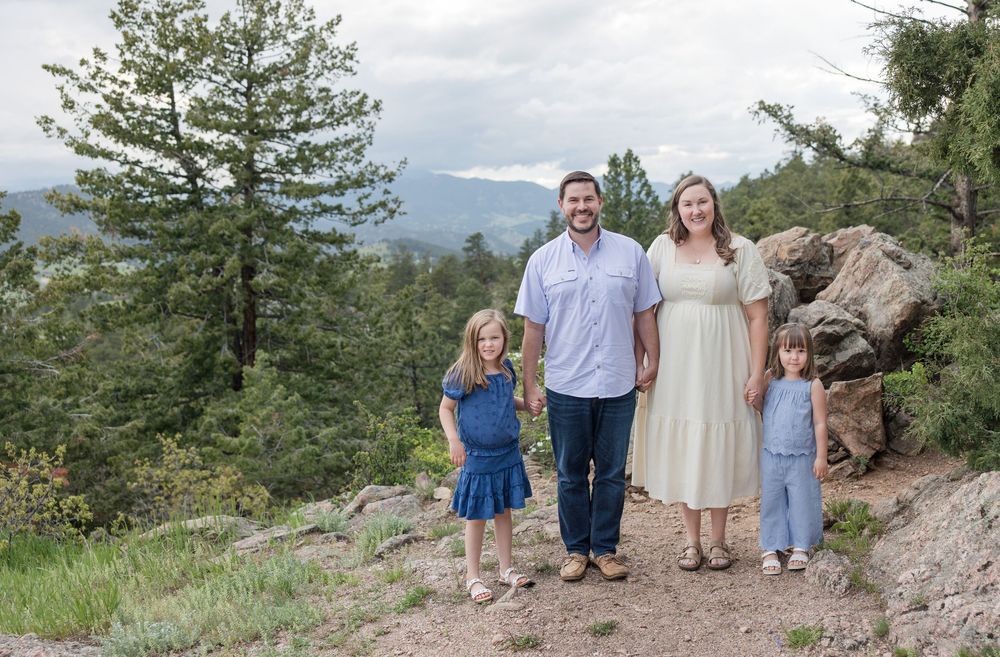 A group stands together on a scenic mountain trail surrounded by evergreen trees and rocky terrain against a cloudy sky.