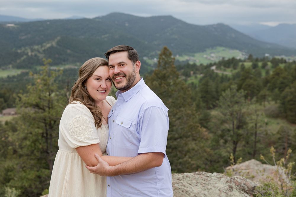 A couple embraces while standing on a scenic mountain overlook with forested peaks in the background.