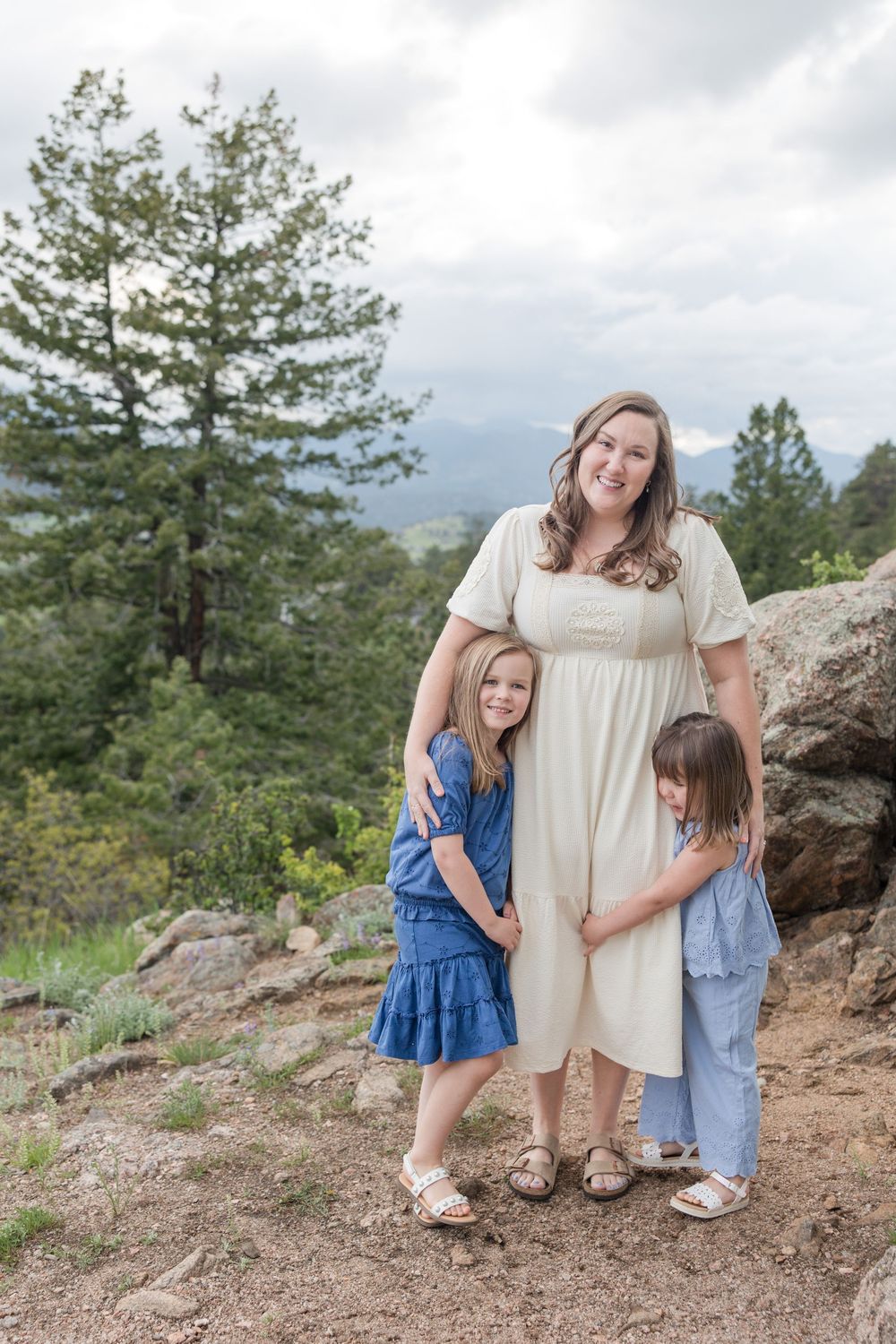 A family poses together on a mountain trail overlooking evergreen trees and rolling hills on a cloudy day.