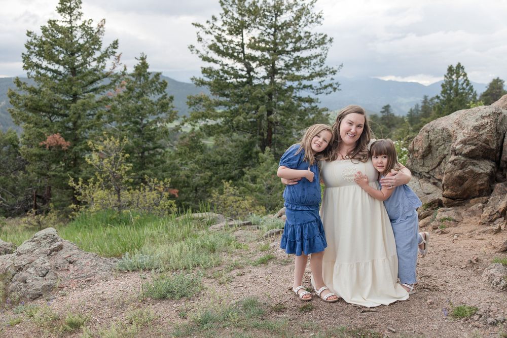 Three figures stand together on a rocky mountain overlook with evergreen trees and mountains in the background.