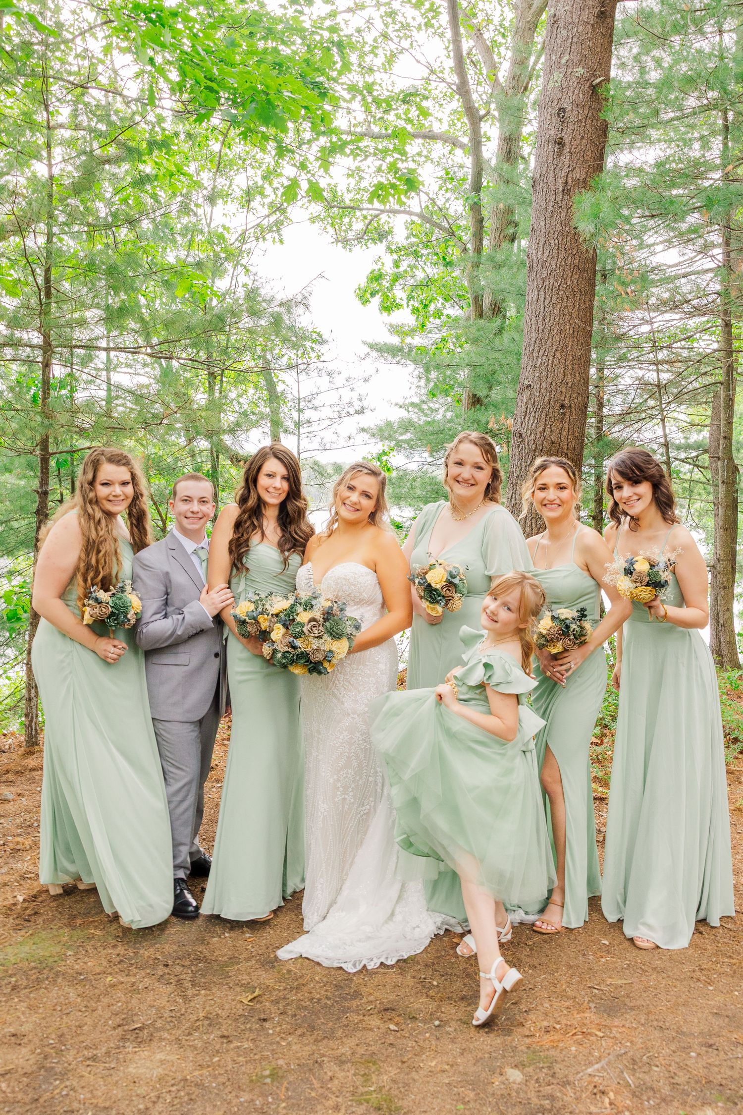 Wedding party wearing sage green bridesmaid dresses pose together in a sunlit forest setting.