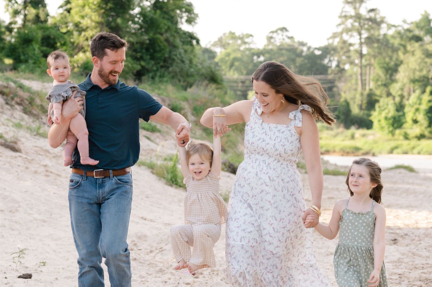 A family joyfully walks together holding hands along a sandy beach path surrounded by green trees on a sunny day.
