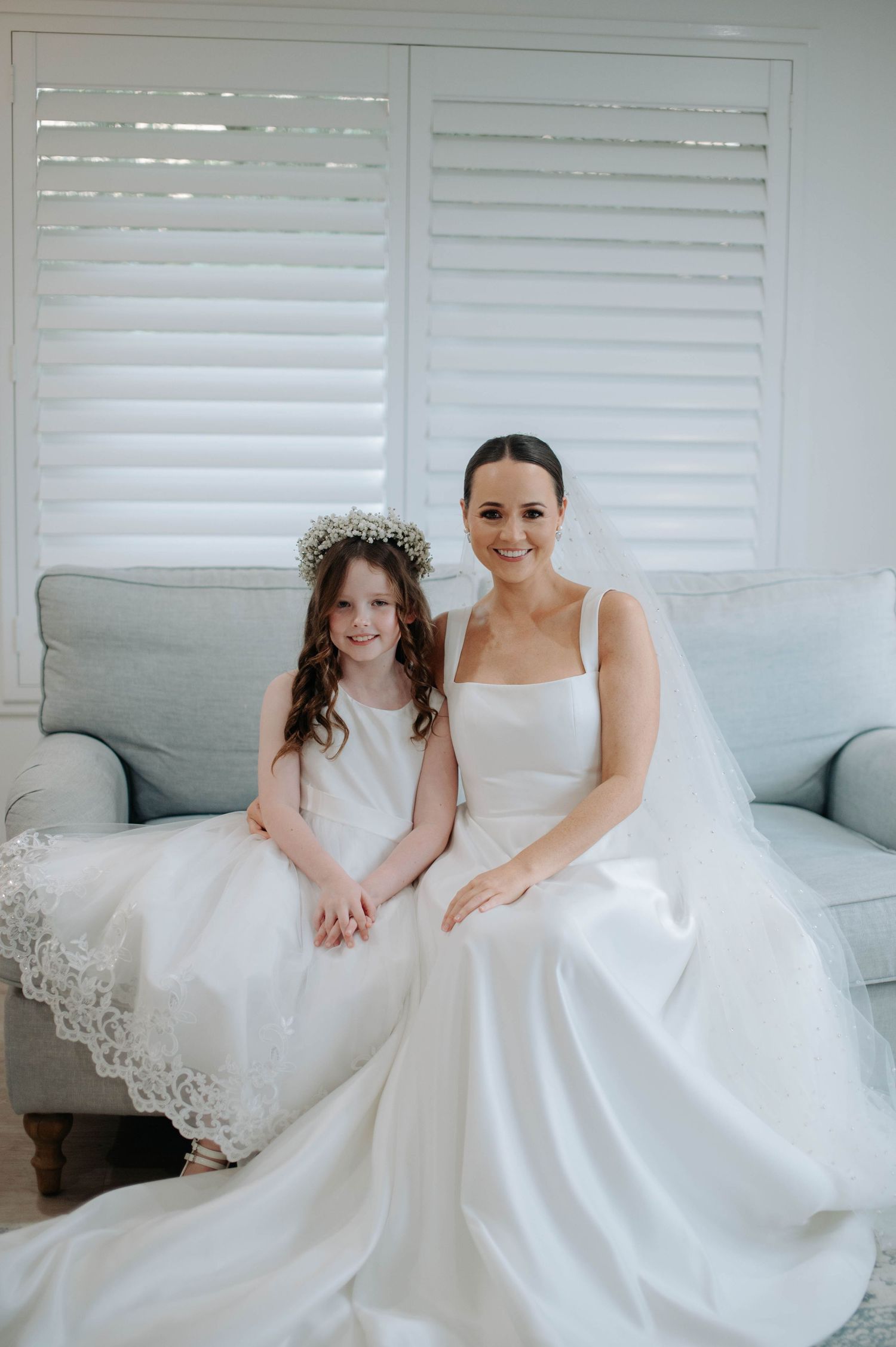 A bride and flower girl wearing matching white dresses sit together on a light gray couch against white shuttered windows.
