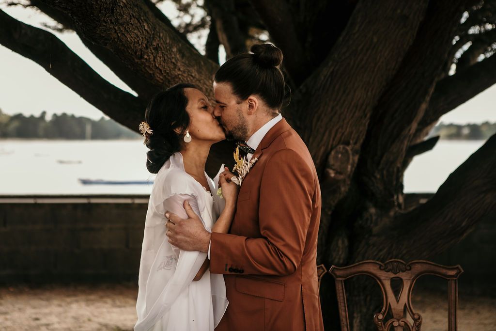 Wedding couple shares intimate moment under a dramatic tree by the water.