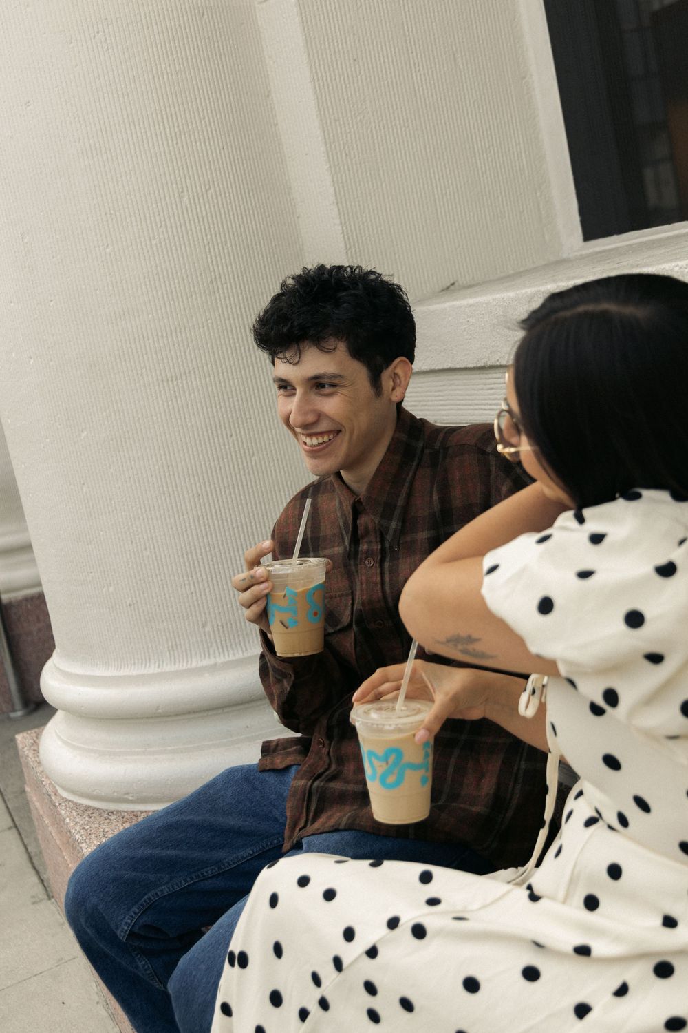 A couple sharing drinks and laughing together while sitting by a white pillar.