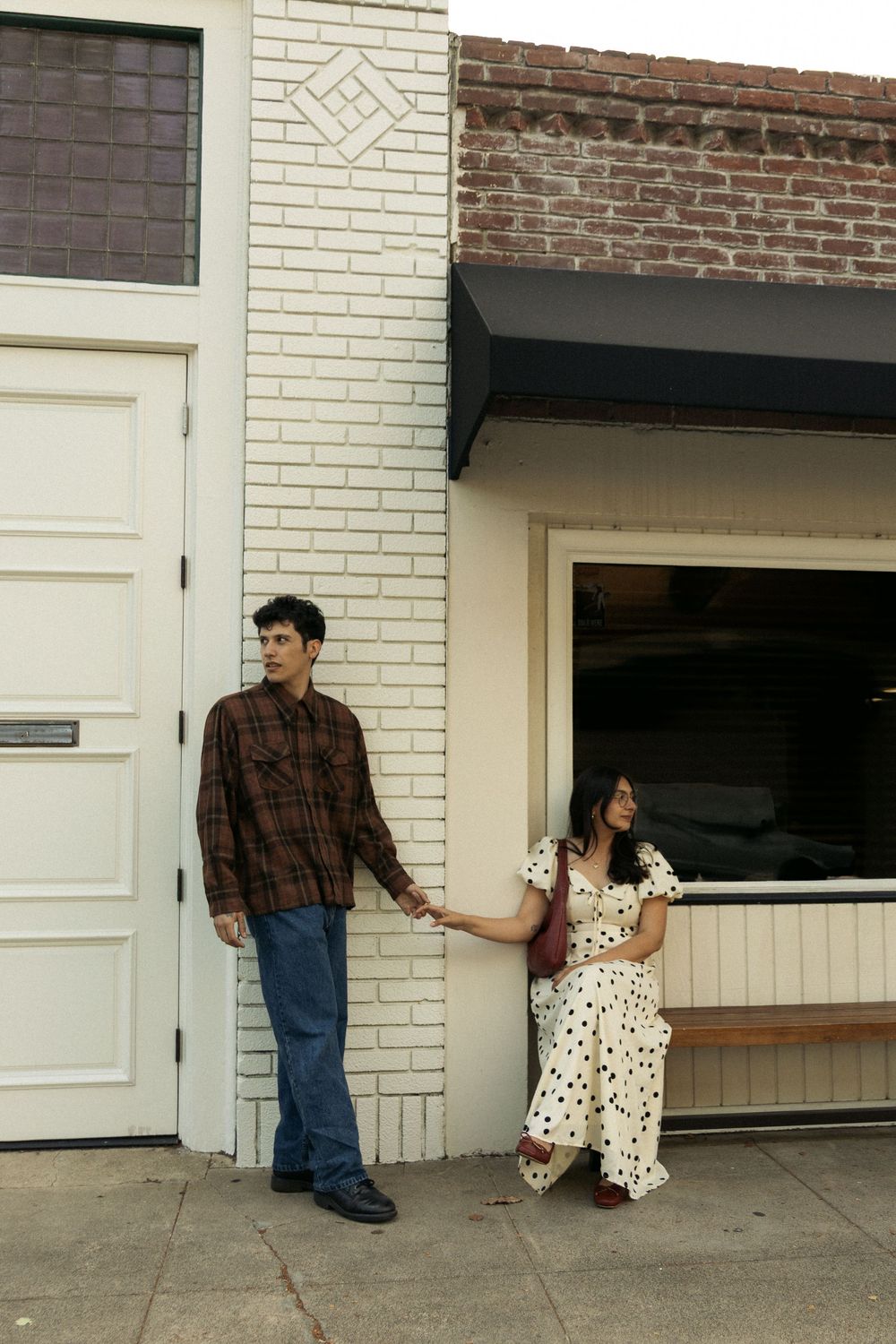 Two people stand casually against a white brick building exterior with a polka dot dress and plaid shirt.