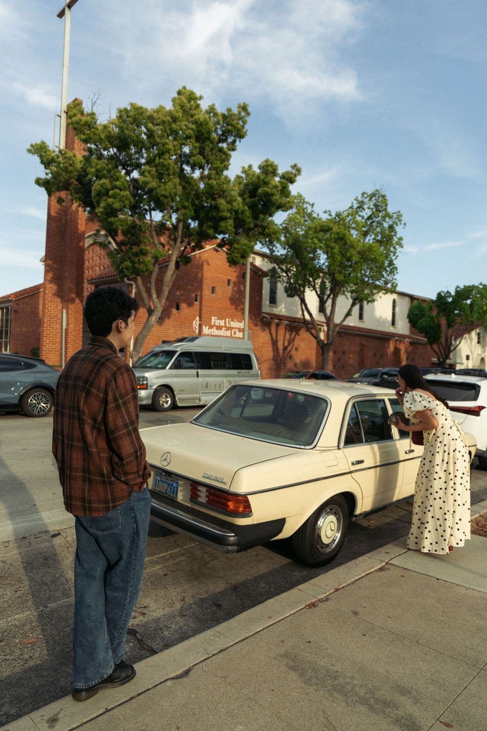 Couple stands beside vintage Mercedes on sunny street with brick buildings and trees.