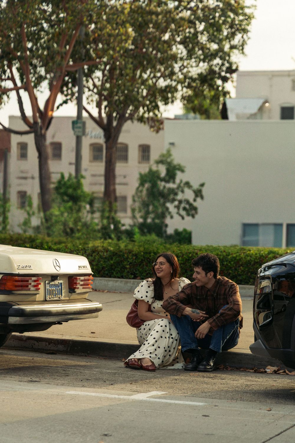 A couple sits on a curb together in vintage-style clothing against an urban backdrop with parked cars.