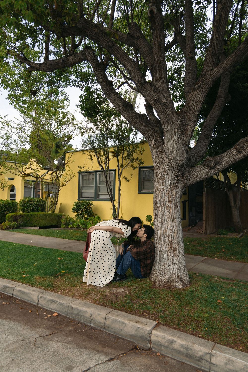 Two people lean against a large tree trunk in front of a yellow house on a residential street.