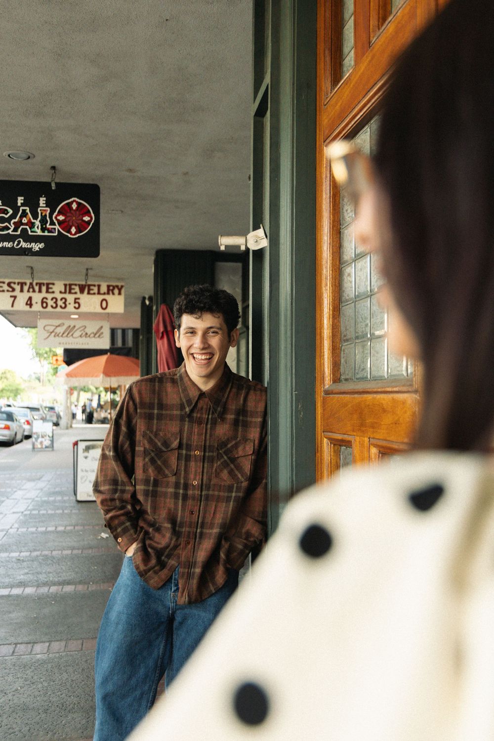 A person wearing a plaid shirt stands smiling outside a store entrance with a vintage storefront sign.