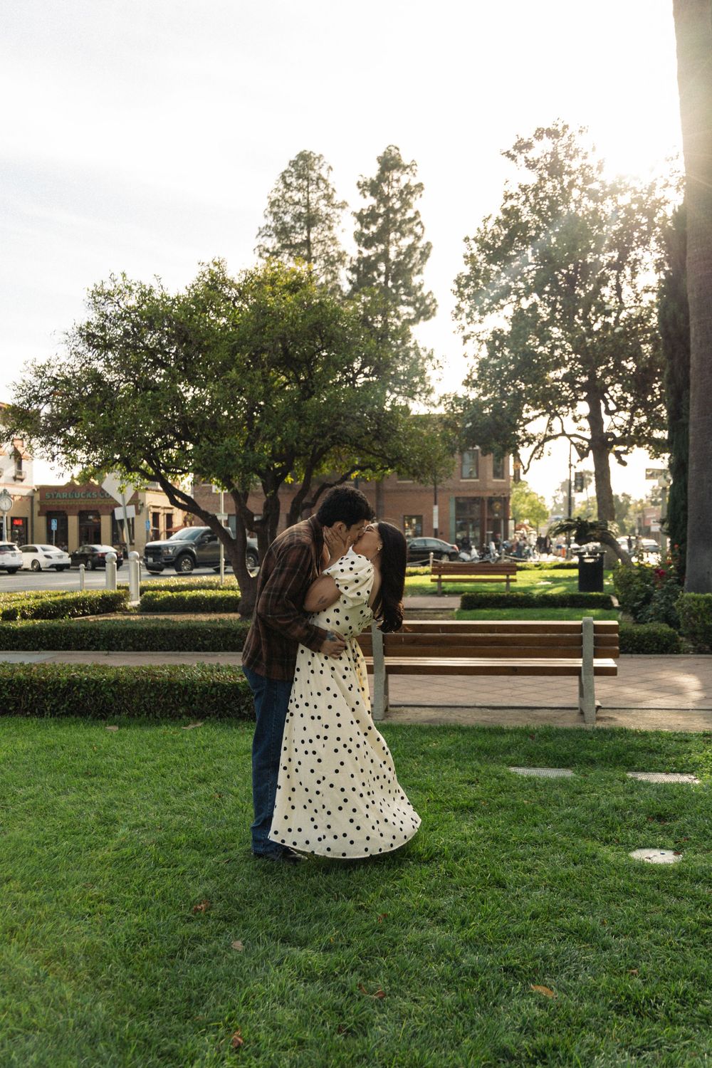 Two people embrace on grass in front of trees, with one wearing a polka dot dress as sunlight filters through.
