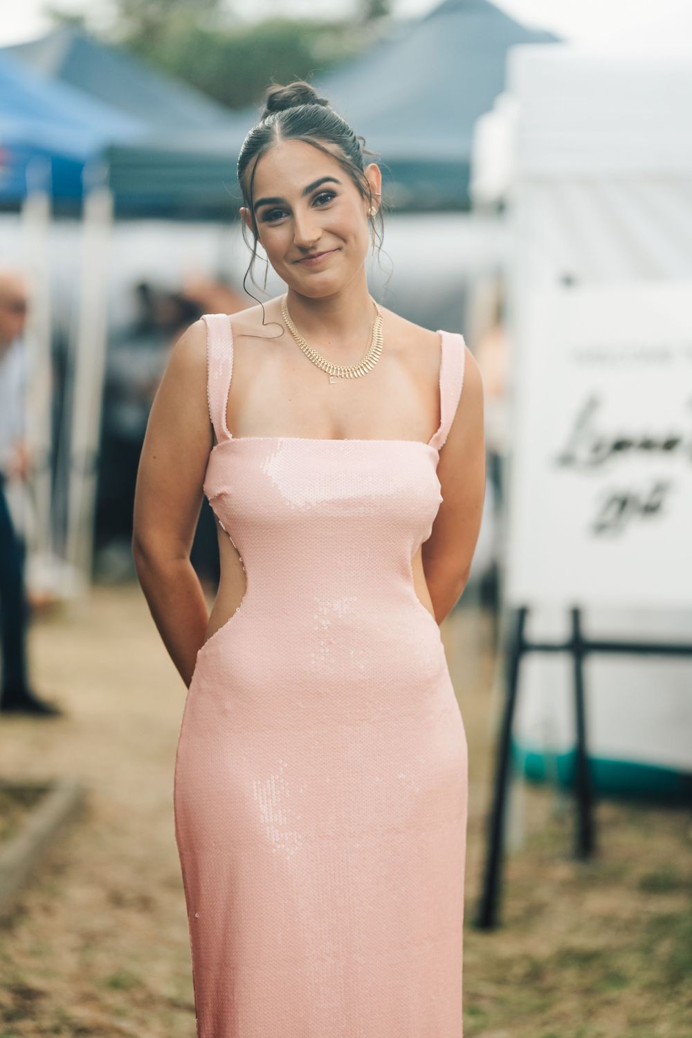 A person in a form-fitting blush pink gown with square neckline and delicate necklace stands outdoors at an event.
