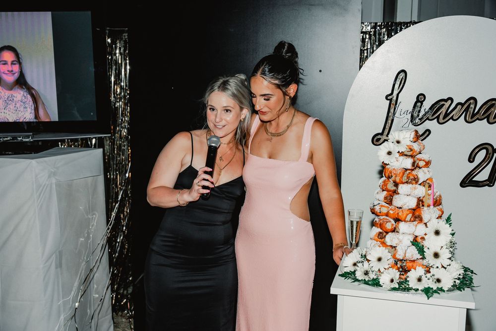 Two women in black and pink evening dresses stand together smiling near an elegant white wedding cake at a reception.