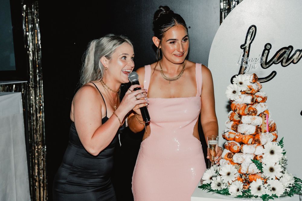 Two women standing by an elegant white wedding cake with orange floral accents at an event.