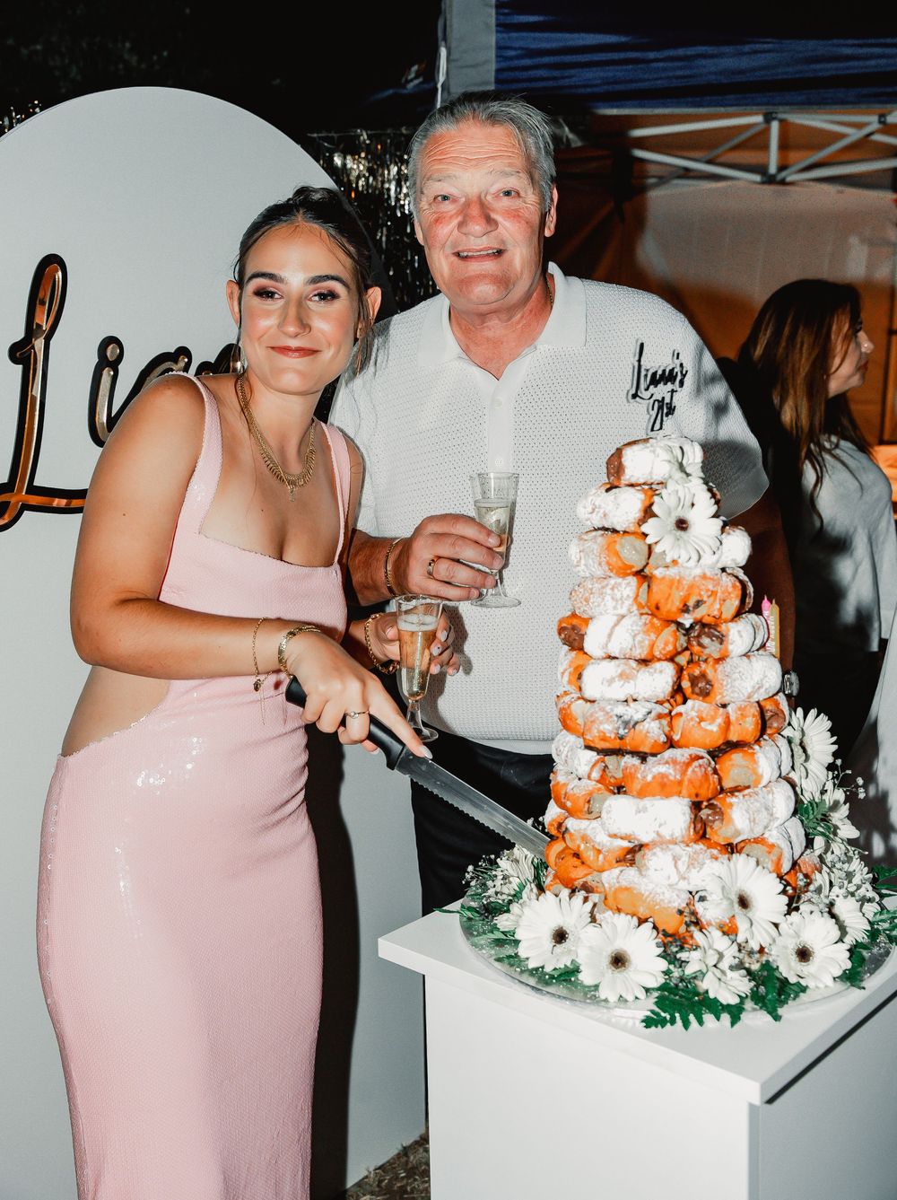 Person in pink dress stands beside a white and orange floral wedding cake during an evening reception.