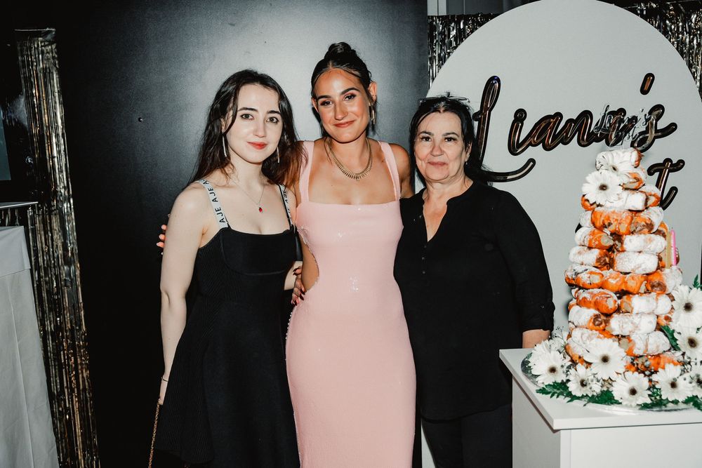 Three people stand together next to an elegant white and coral-colored wedding cake at an event venue.