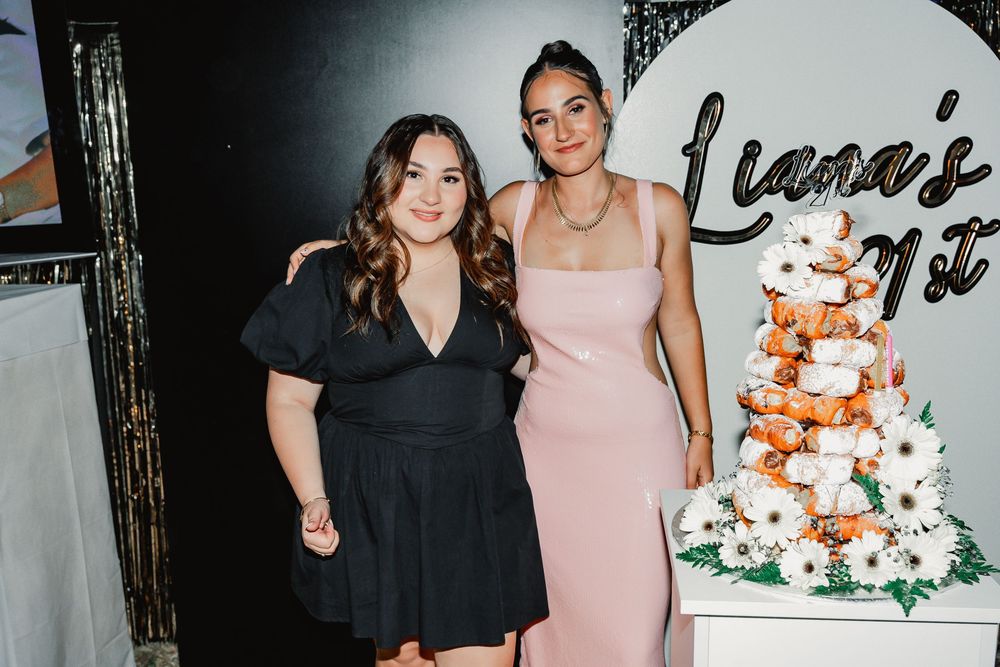 Two friends pose together at a party celebration next to an elegant white floral decorated cake display.